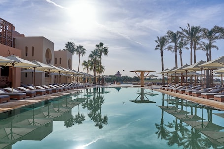 A poolside area with lounge chairs and umbrellas, surrounded by palm trees and a clear sky.