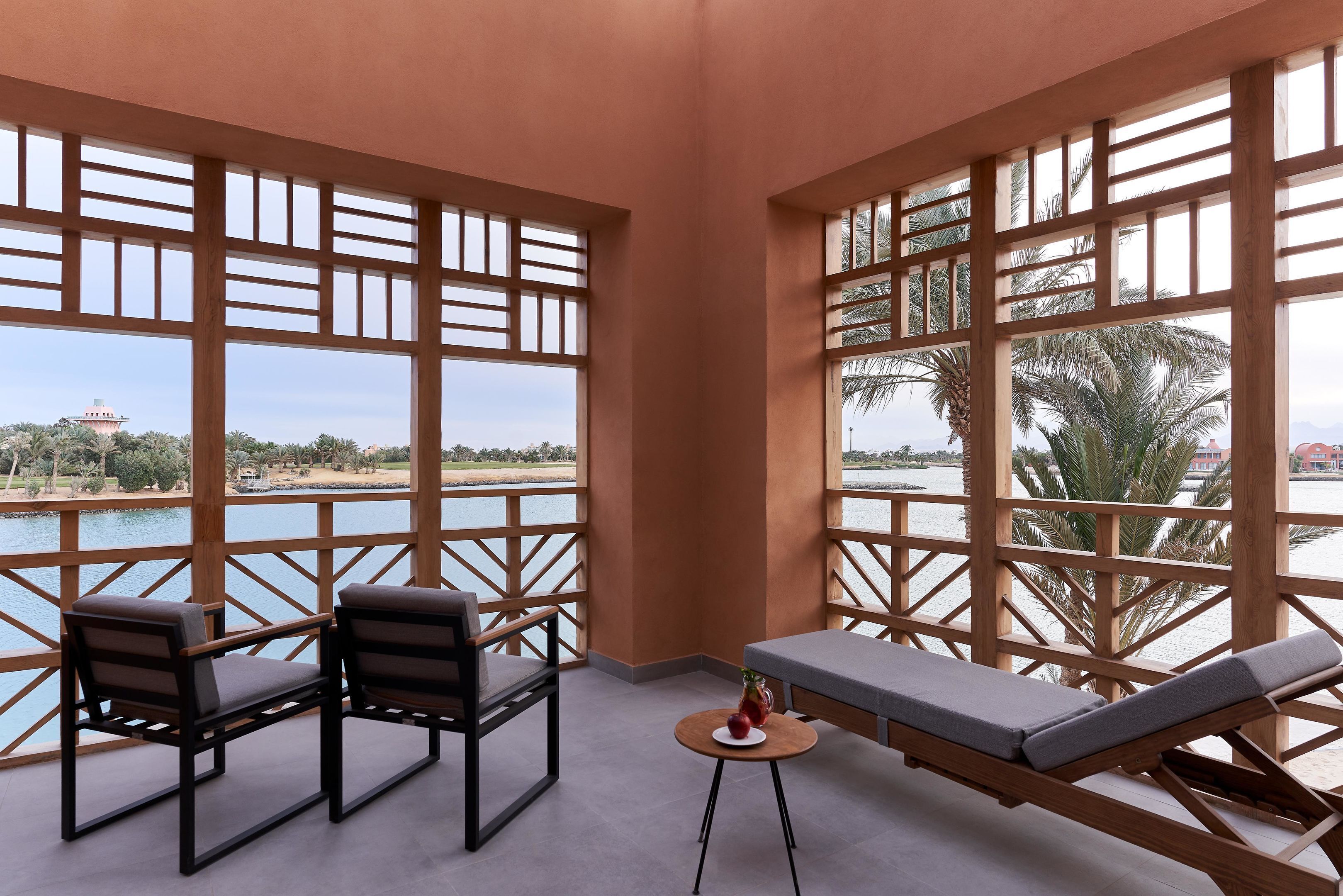 A balcony with wooden lattice panels, two chairs, a small table with a fruit bowl, and a lounge chair.