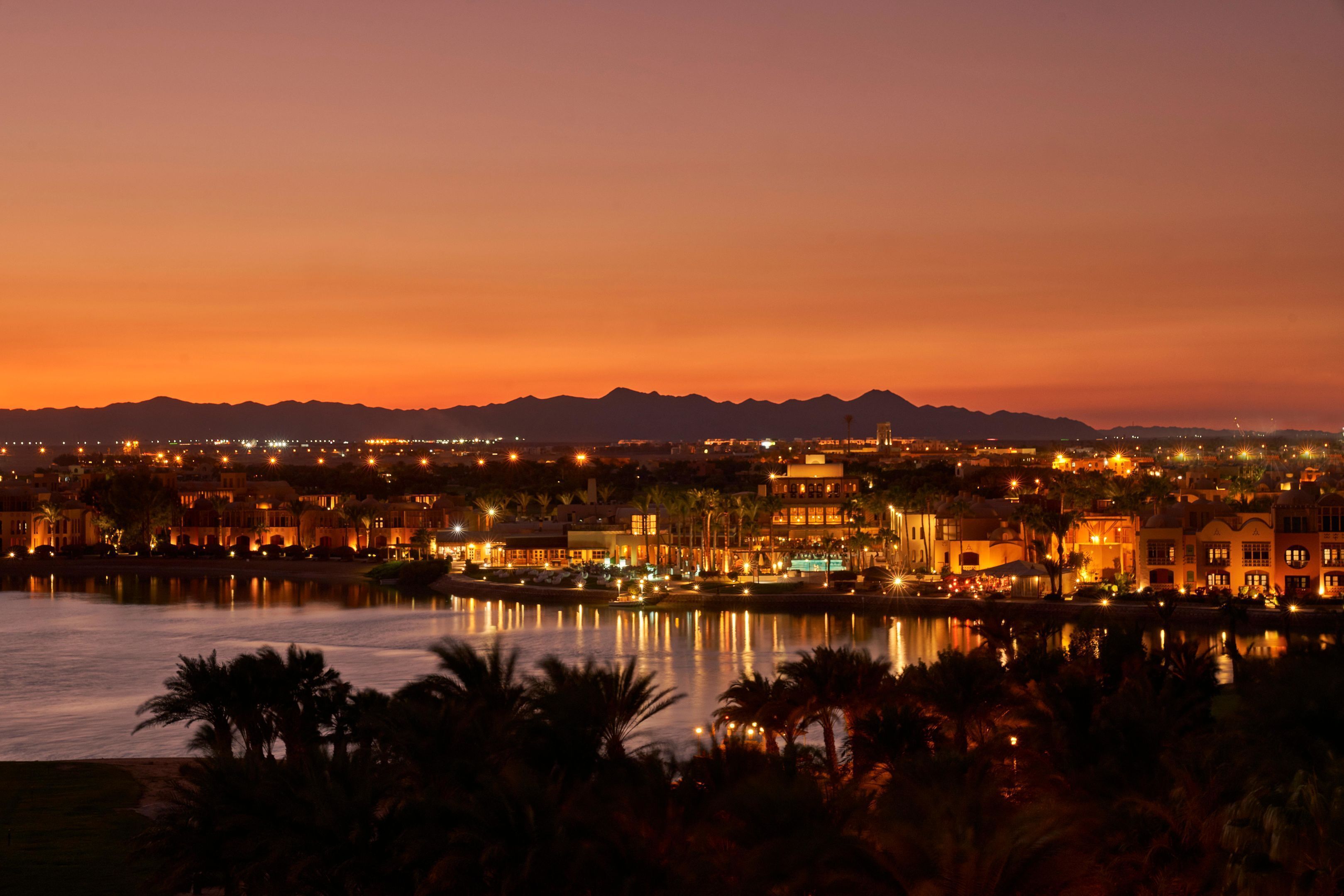 A nighttime cityscape with illuminated buildings, a body of water, and mountains in the background.