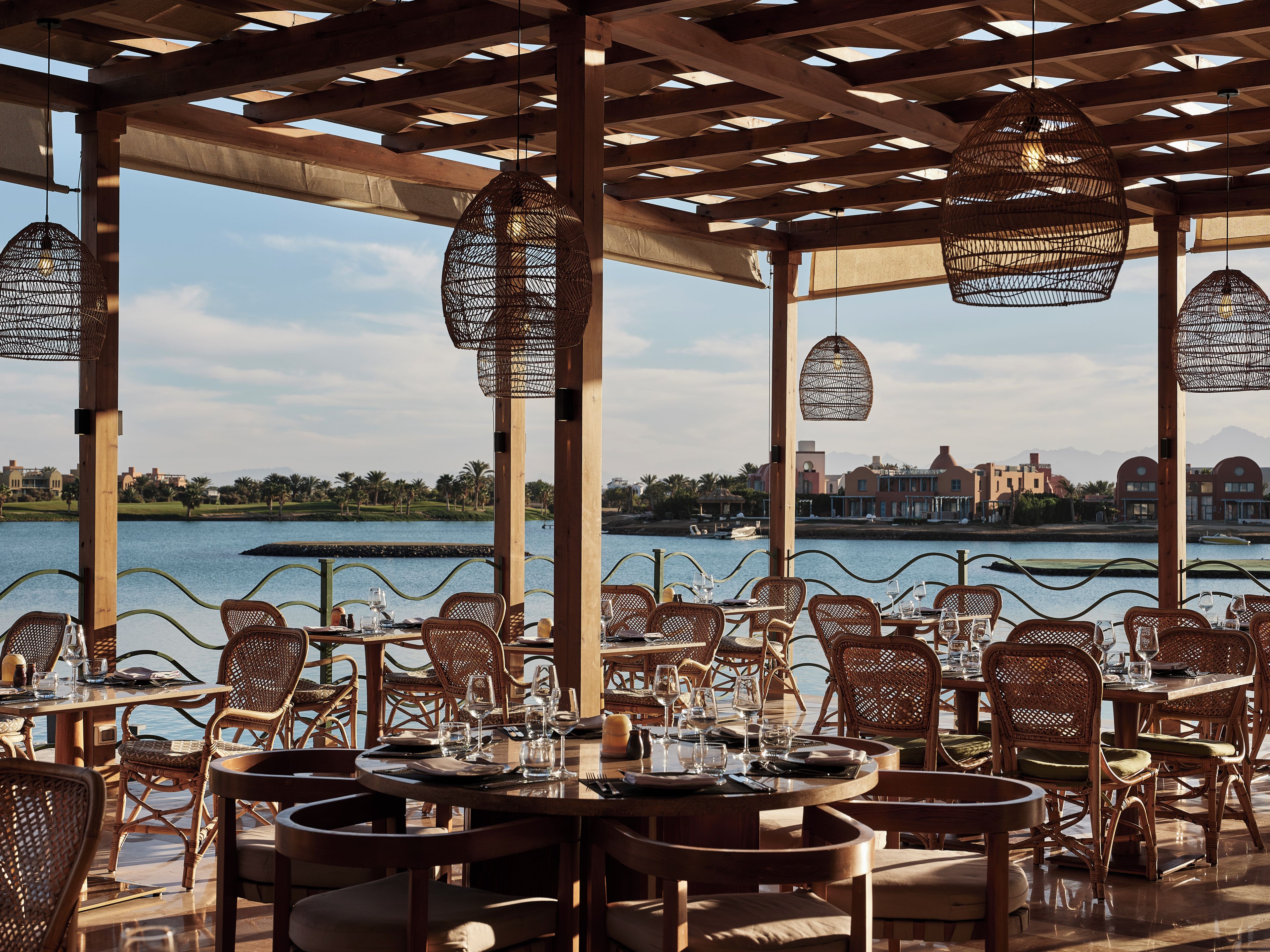 An outdoor dining area with tables and chairs, overlooking a body of water and a distant cityscape.