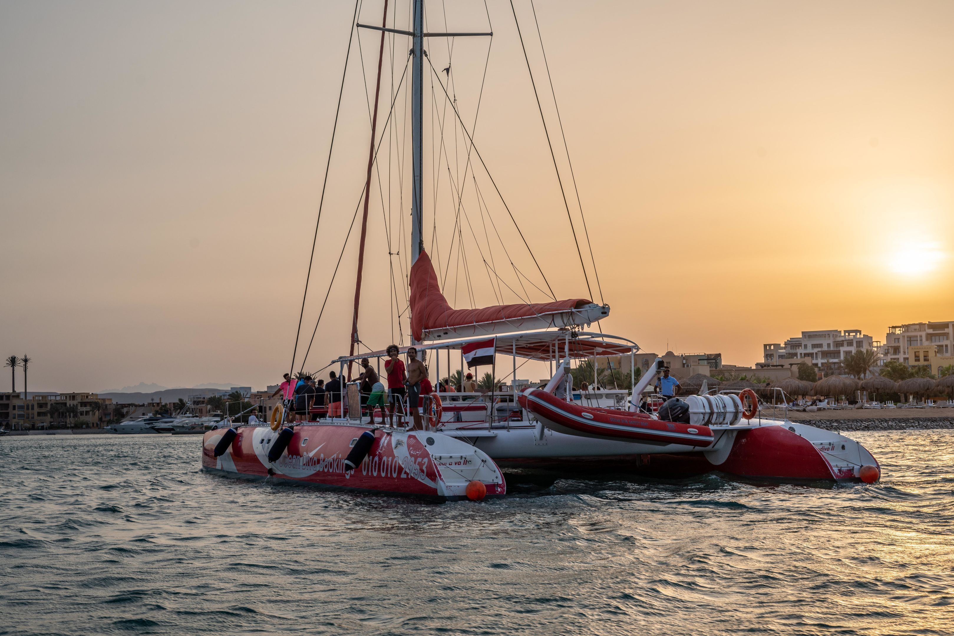 Un bateau rouge et blanc transportant des passagers est amarré près d’une promenade en bord de mer, avec des bâtiments en arrière-plan.
