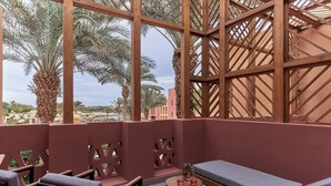 A balcony with a wooden lattice structure, a cushioned bench, and a small table with a vase and two glasses.