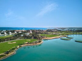 Vista aérea de un complejo turístico costero con campos de golf, cuerpos de agua y villas frente a la playa.