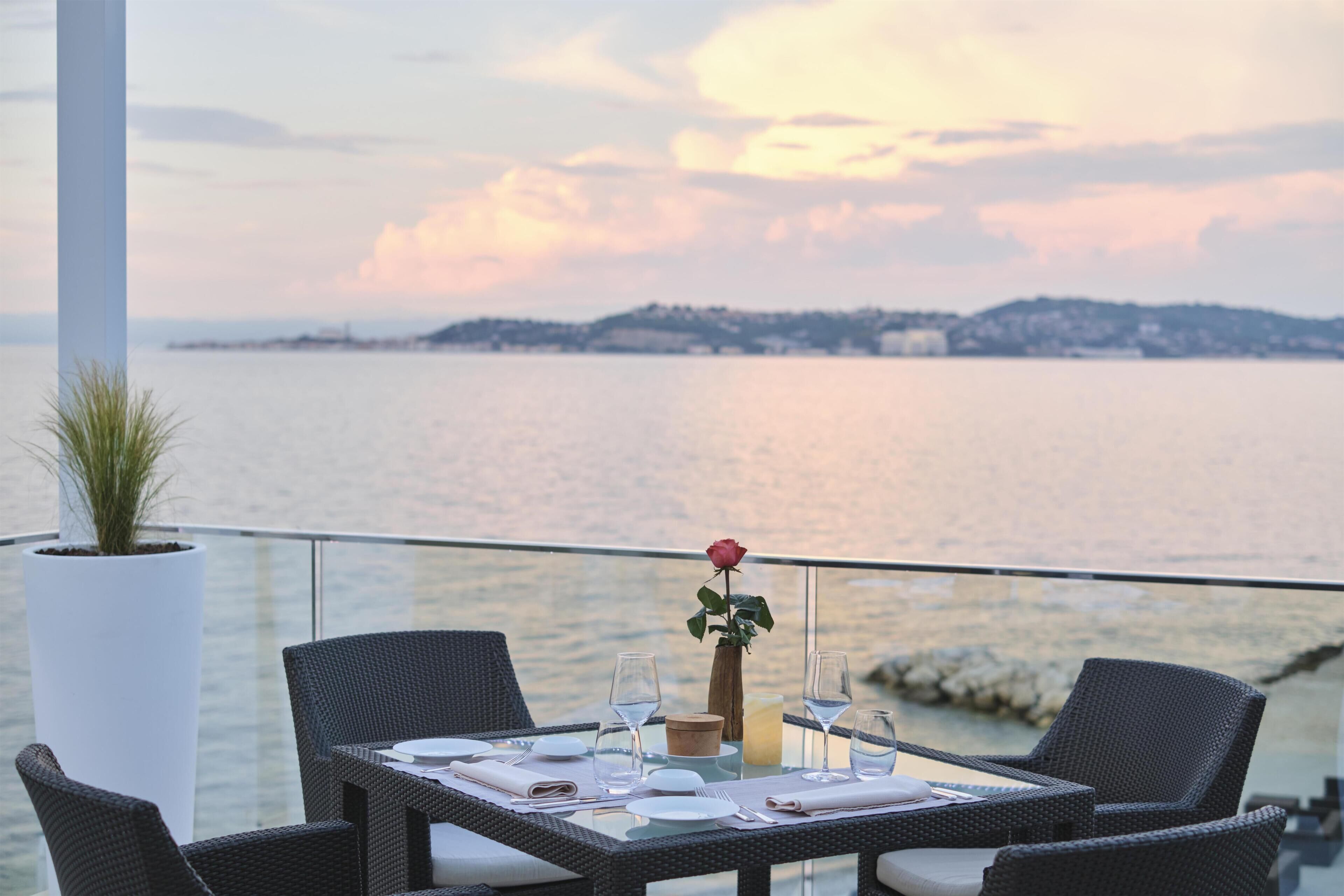 Una mesa de comedor dispuesta con platos, vasos y un jarrón con una sola rosa, con vistas a un cuerpo de agua y una orilla lejana.