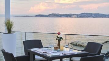Una mesa de comedor dispuesta con platos, vasos y un jarrón con una sola rosa, con vistas a un cuerpo de agua y una orilla lejana.