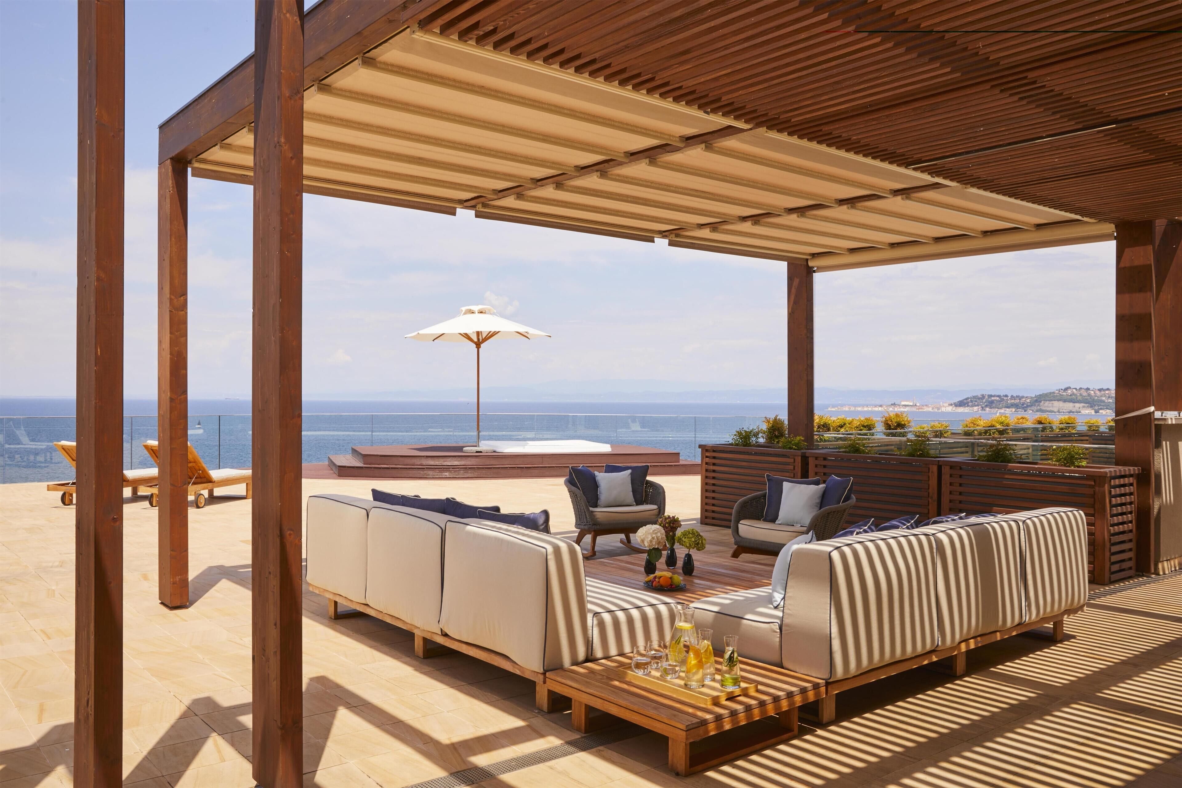 Un área de estar al aire libre con techo, con vistas al mar, amueblada con sofás y sillones, y una mesa con bebidas.