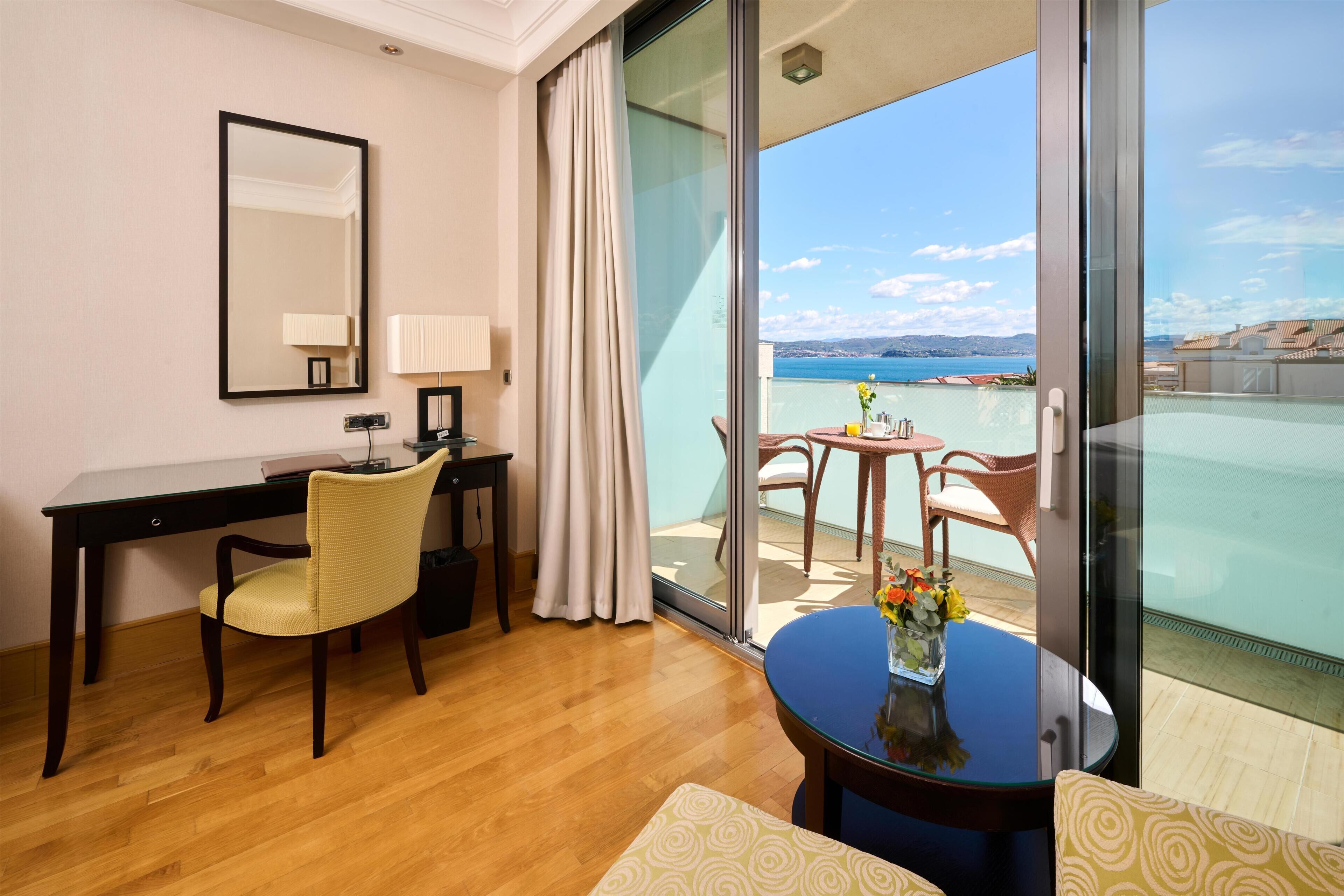 Une chambre d’hôtel avec un bureau en bois, une chaise, une petite table sur laquelle est posé un vase de fleurs, un grand miroir et une porte-fenêtre coulissante donnant sur un balcon avec vue sur la mer.
