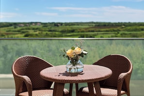 A wicker table with two chairs and a vase of flowers, set against a scenic view of a lake and greenery.
