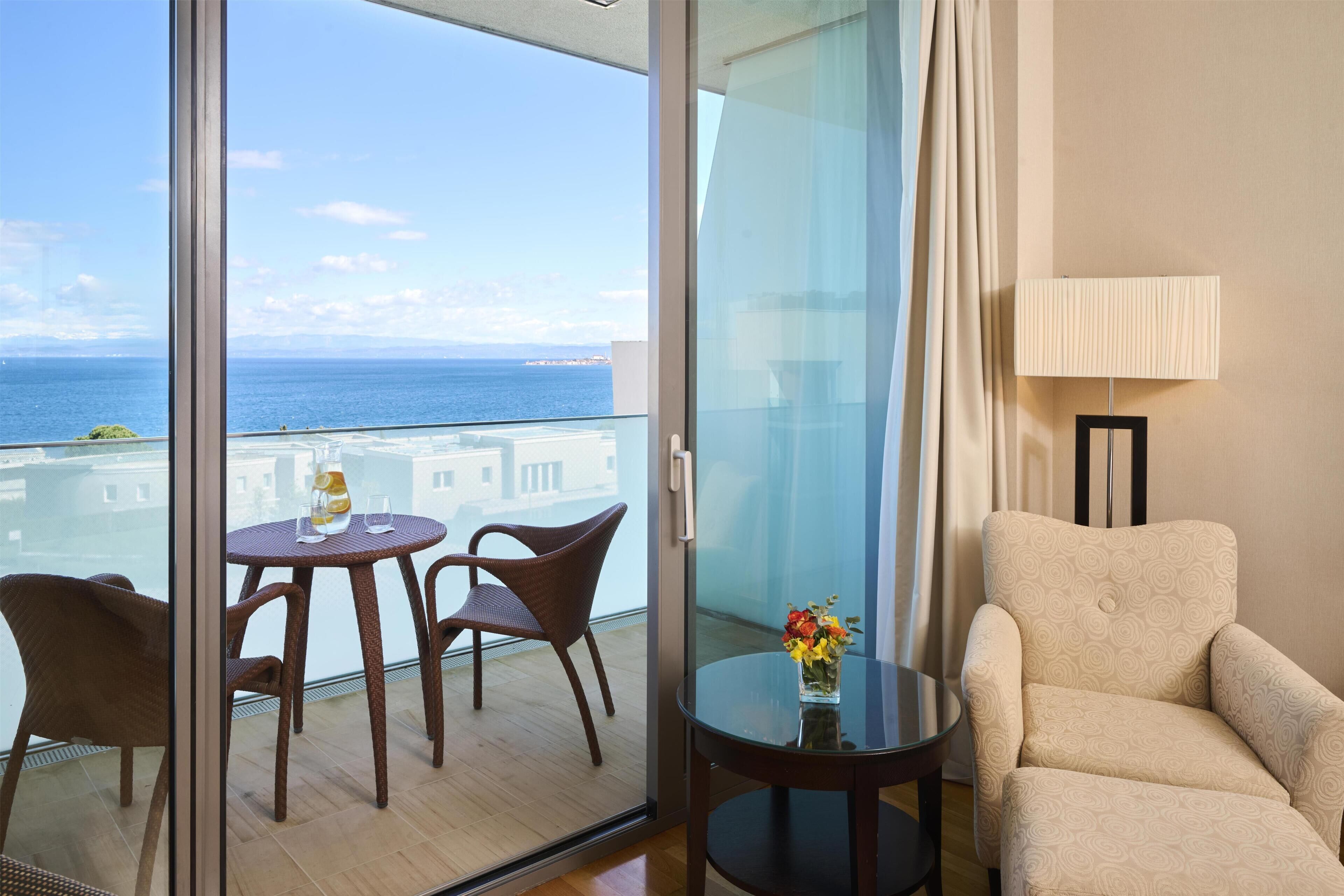 A balcony with a view of the sea, a table with chairs, a vase with flowers, and a lamp.