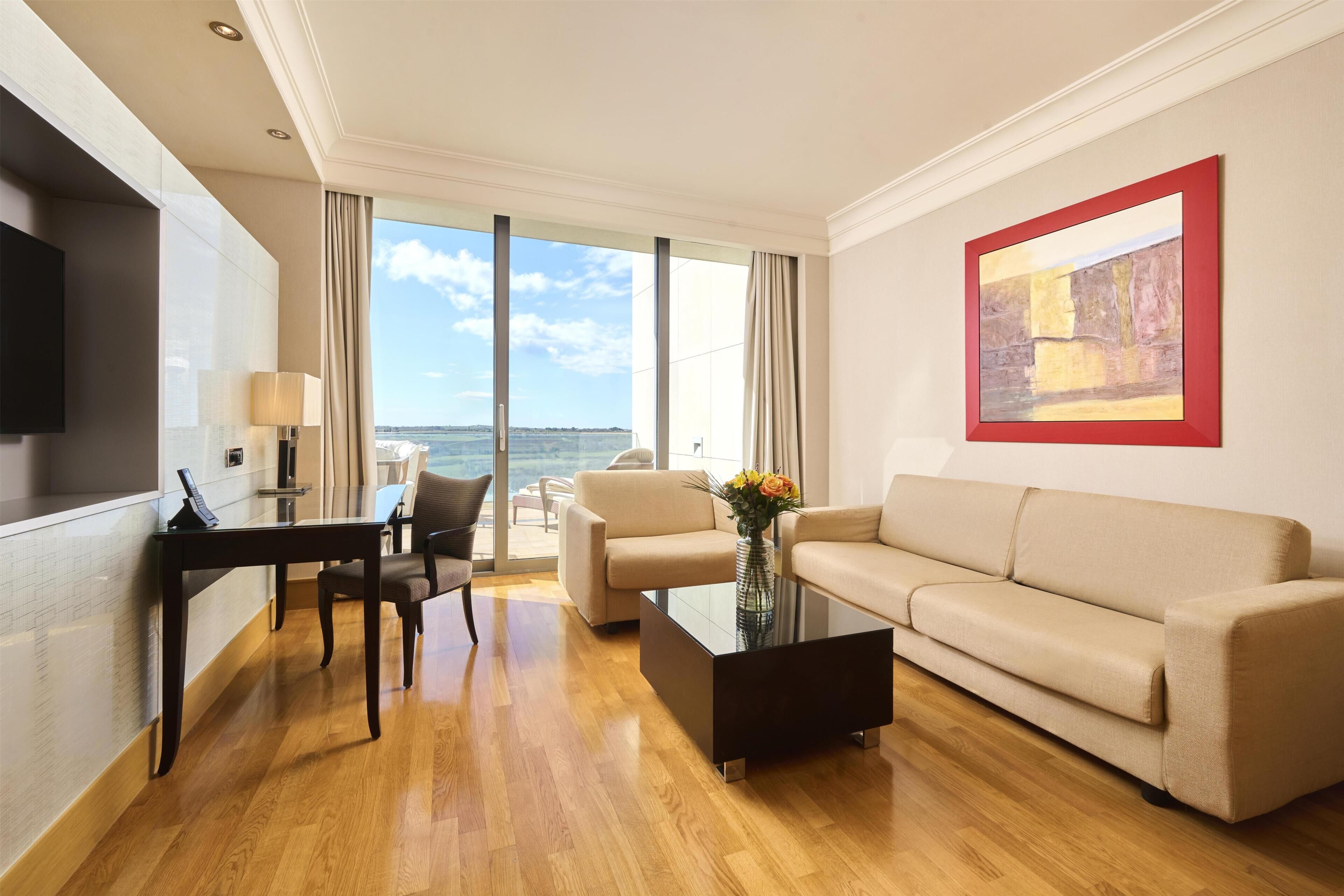 A modern hotel room with a sofa, a coffee table, a TV, and a large window offering a view of the sea.