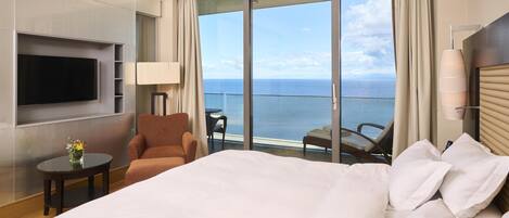 Habitación de hotel con una cama grande, un televisor montado en la pared, una silla, una mesita con un jarrón y un balcón con vistas al mar.