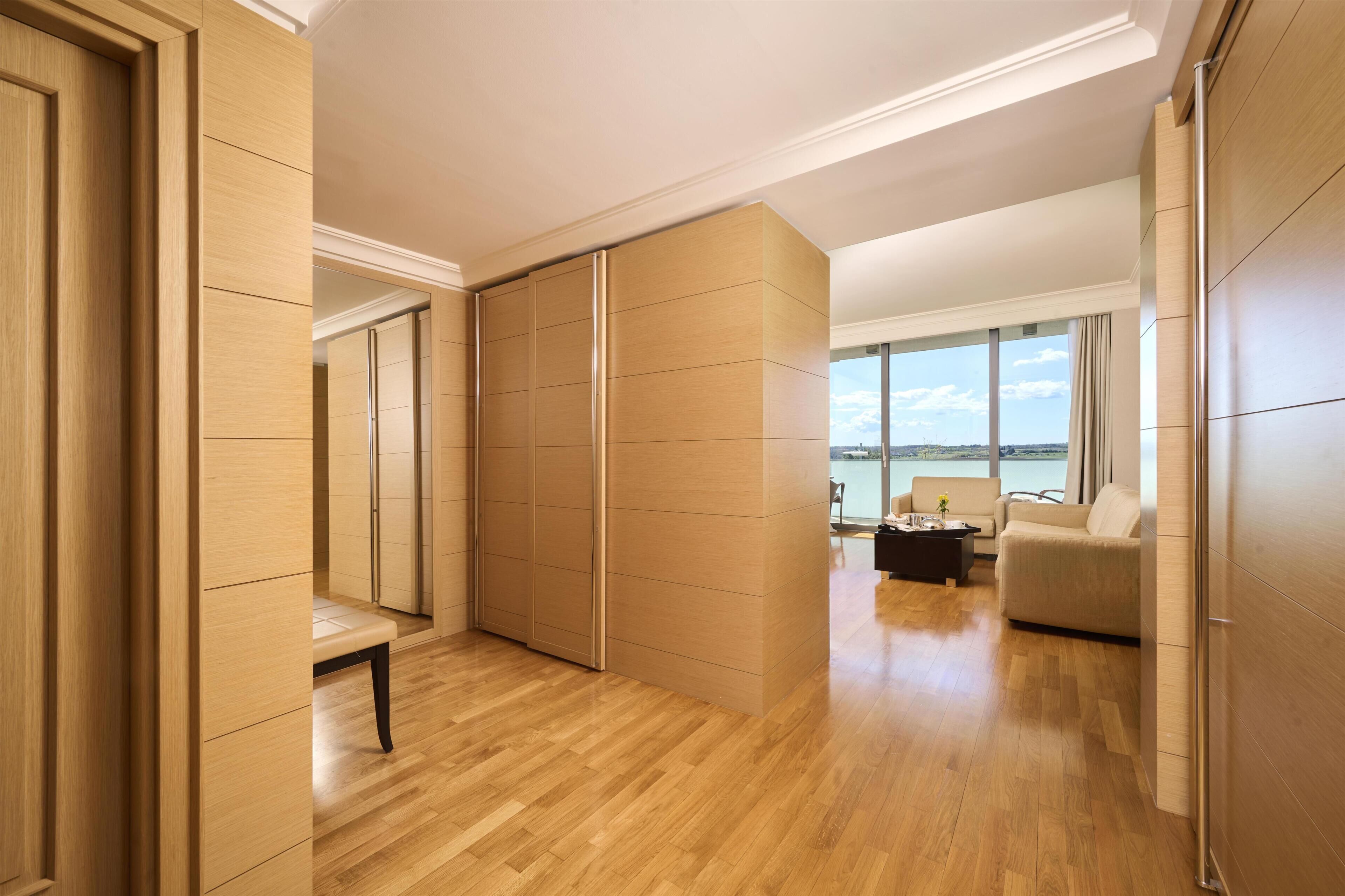 Une chambre d’hôtel moderne avec un plancher en bois, une armoire à portes coulissantes, un coin salon avec un canapé et une petite table, et une grande fenêtre donnant sur la mer.