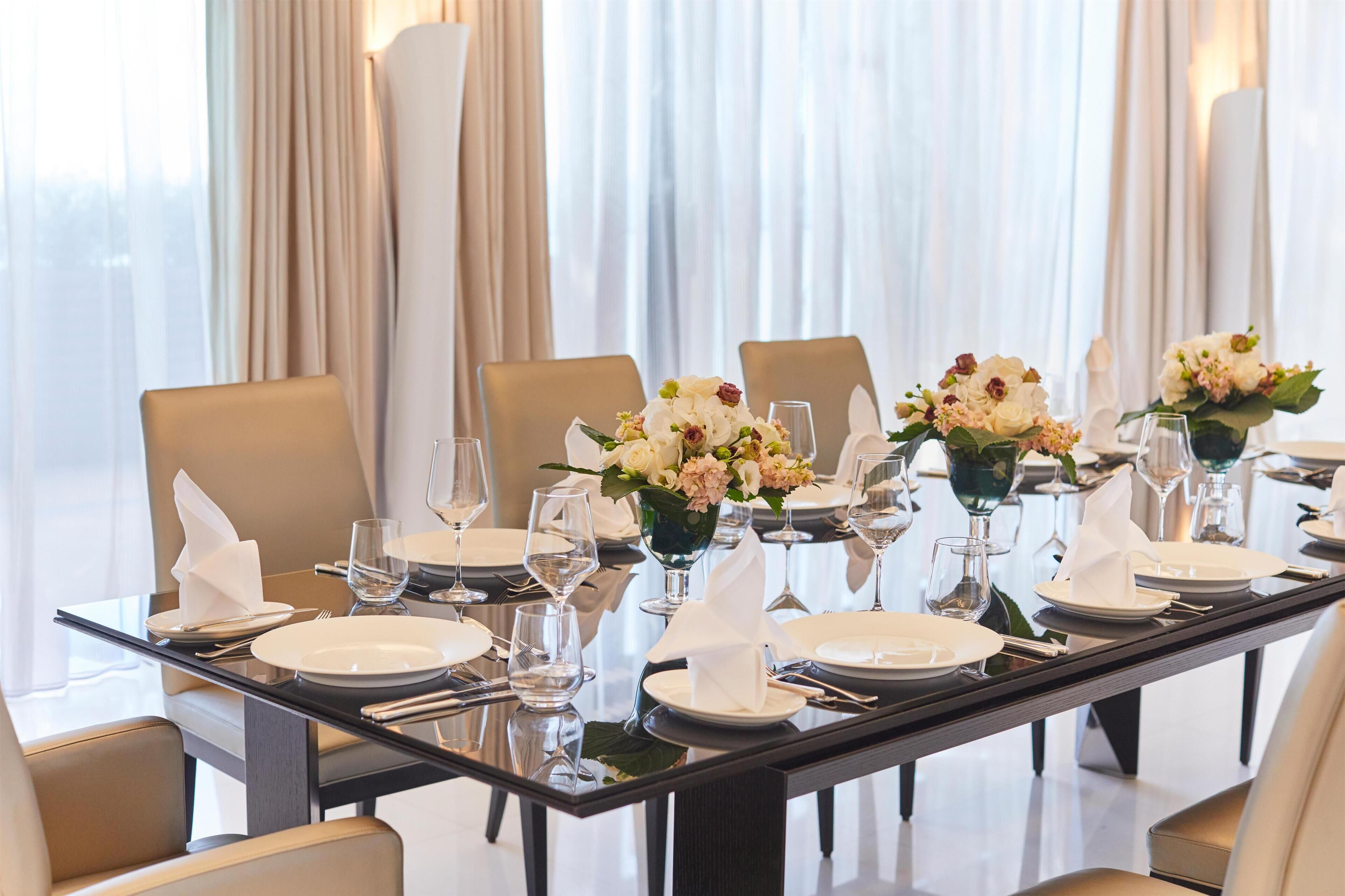 Una mesa de comedor dispuesta para una comida formal, con platos blancos, servilletas dobladas y centros de mesa con flores.