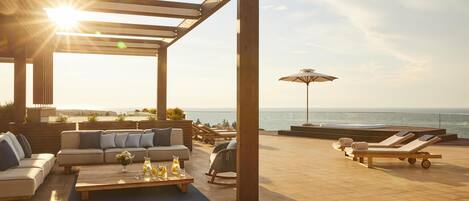 Un espacio lounge al aire libre iluminado por el sol, con una pérgola de madera, un conjunto de sofás, una mesa de centro, sillas de relax y vistas al océano.