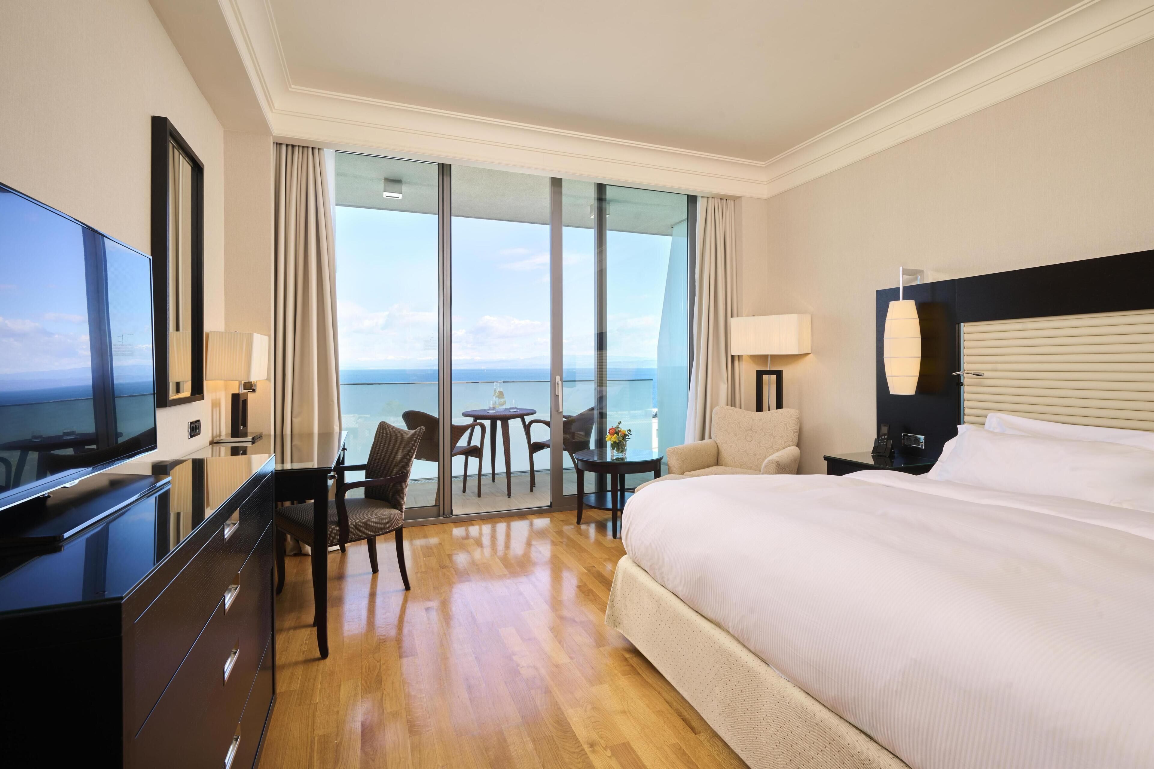 A hotel room with a large bed, a TV, a desk with a chair, a small table with a lamp, and a view of the sea through large windows.