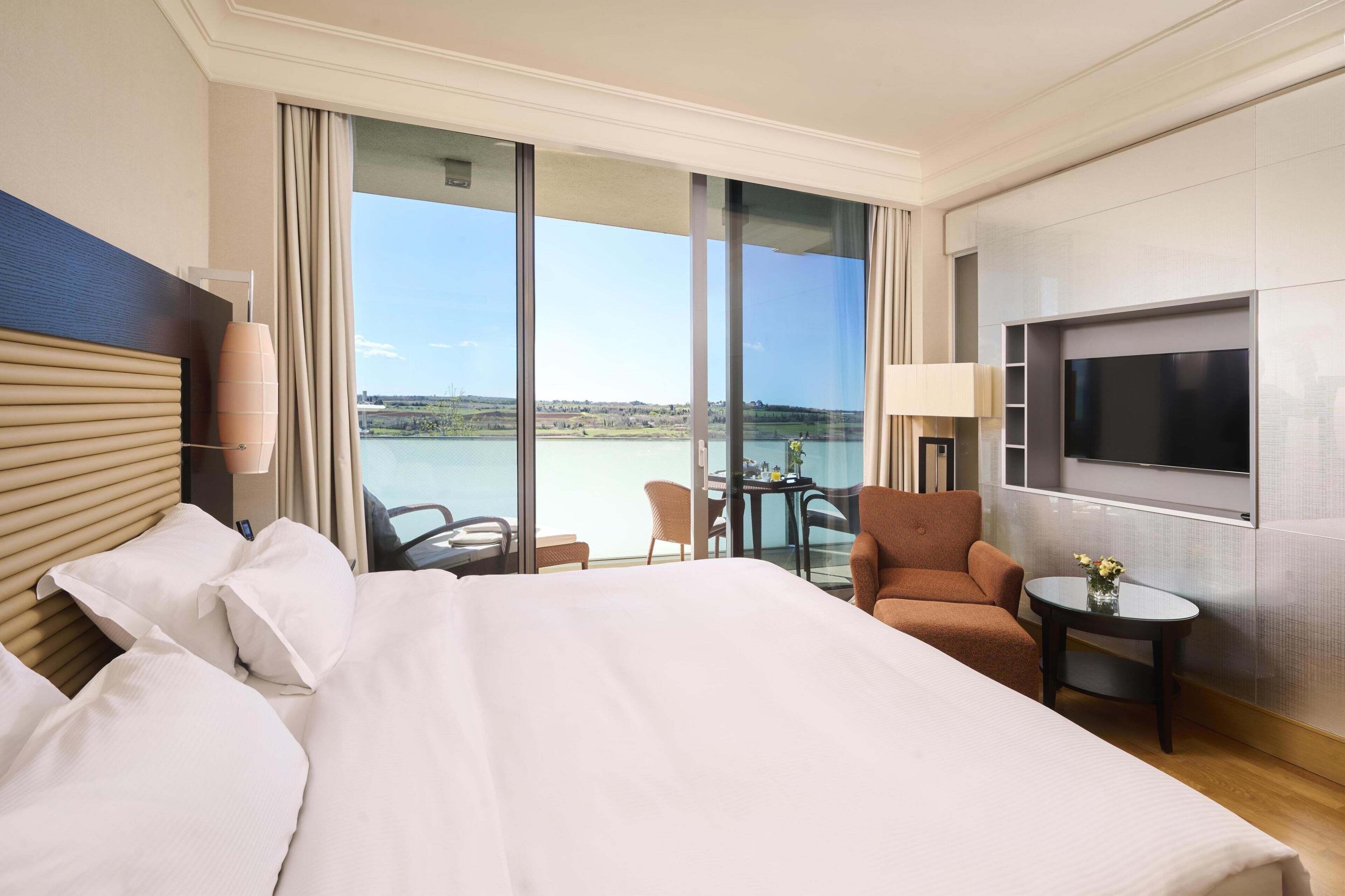 A hotel room with a large bed, a TV, a small table with a vase, and a chair.