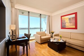 Una habitación de hotel moderna con una puerta de cristal que da a un balcón, un sofá, una mesa de centro y un escritorio con una lámpara.