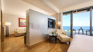 A modern hotel room with a large bed, a sofa, a chair, a TV, and a balcony with a view of the sea.