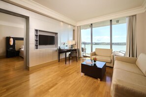 Ett modernt hotellrum med trägolv, en platt-TV som är monterad på väggen, ett skrivbord med en stol, en soffa och ett stort fönster med utsikt över en sjö eller ett annat vattendrag.