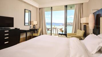 Ein Hotelzimmer mit einem großen Bett, einem Schreibtisch mit Stuhl, einem Fernseher und Meerblick durch große Fenster.
