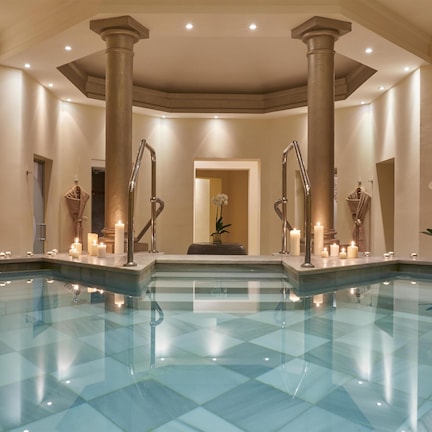 A spacious indoor swimming pool with clear water, surrounded by columns and potted plants.