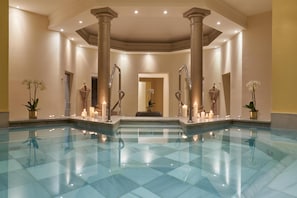 A spacious indoor swimming pool with clear water, surrounded by columns and potted plants.