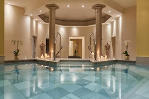 A spacious indoor swimming pool with clear water, surrounded by columns and potted plants.