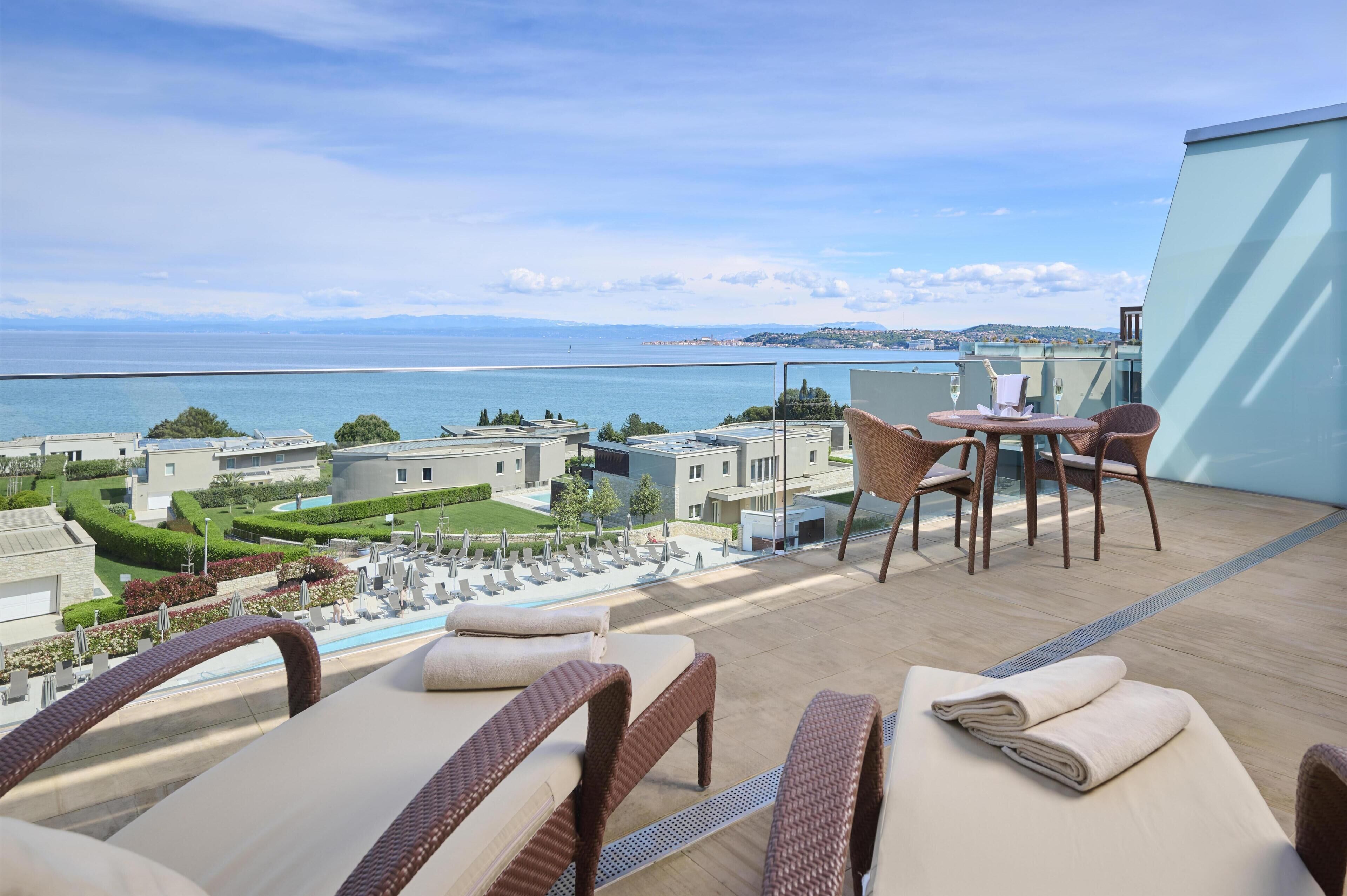 A balcony with wicker chairs and a table, overlooking a pool and a view of the sea and buildings.