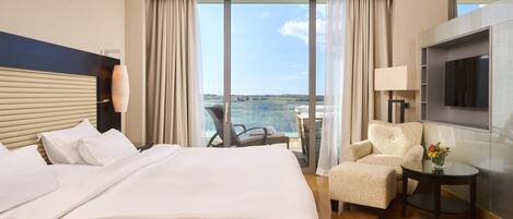 A hotel room with a large bed, a chair, a small table, a TV, and a view of the sea.