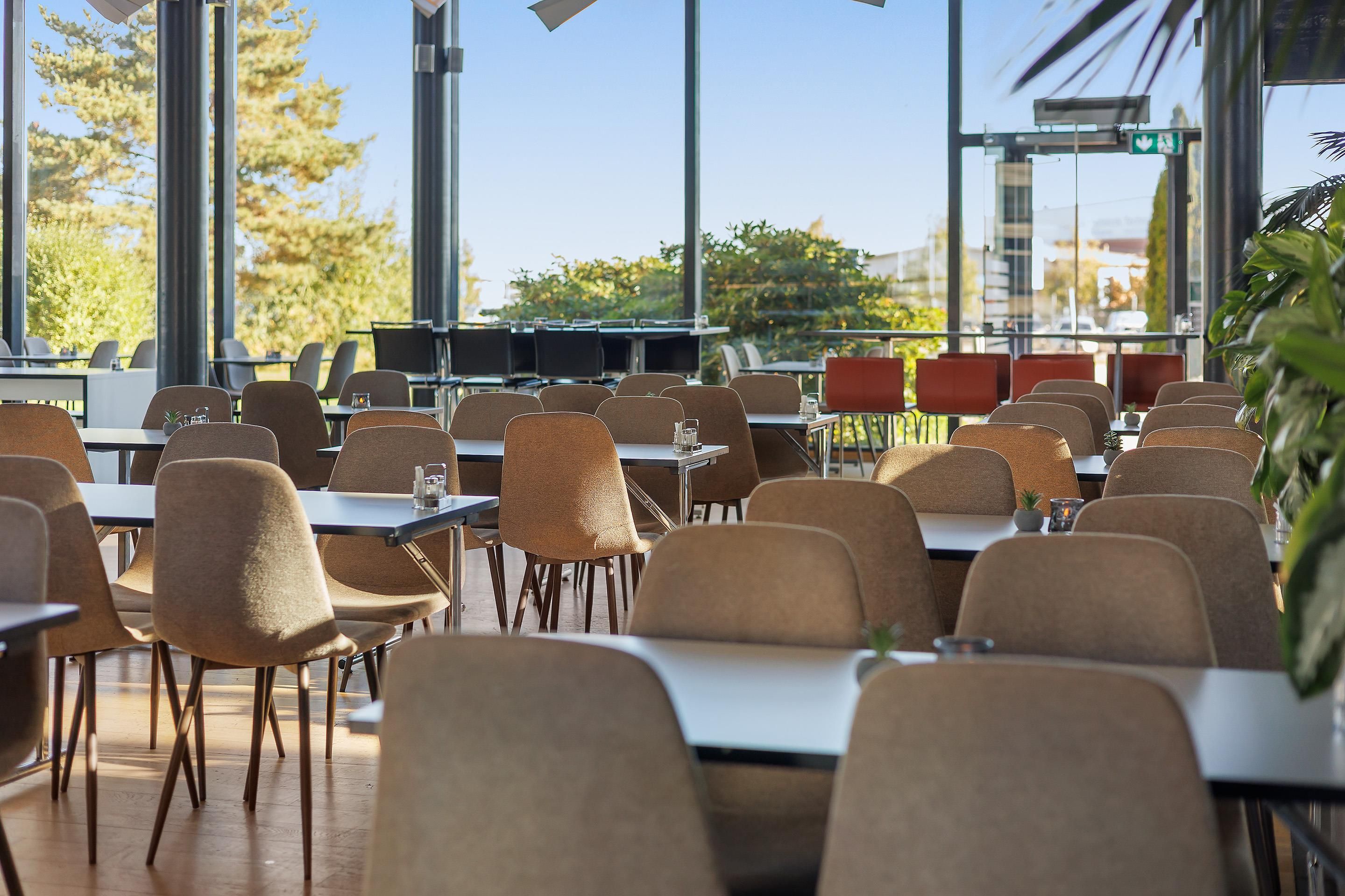 Een eetruimte met tafels en stoelen, grote ramen en uitzicht op bomen en gebouwen.