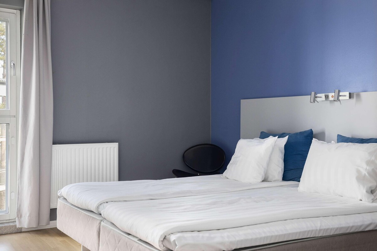 A bedroom with a bed, a radiator, a window with curtains, and a wall with a blue accent.