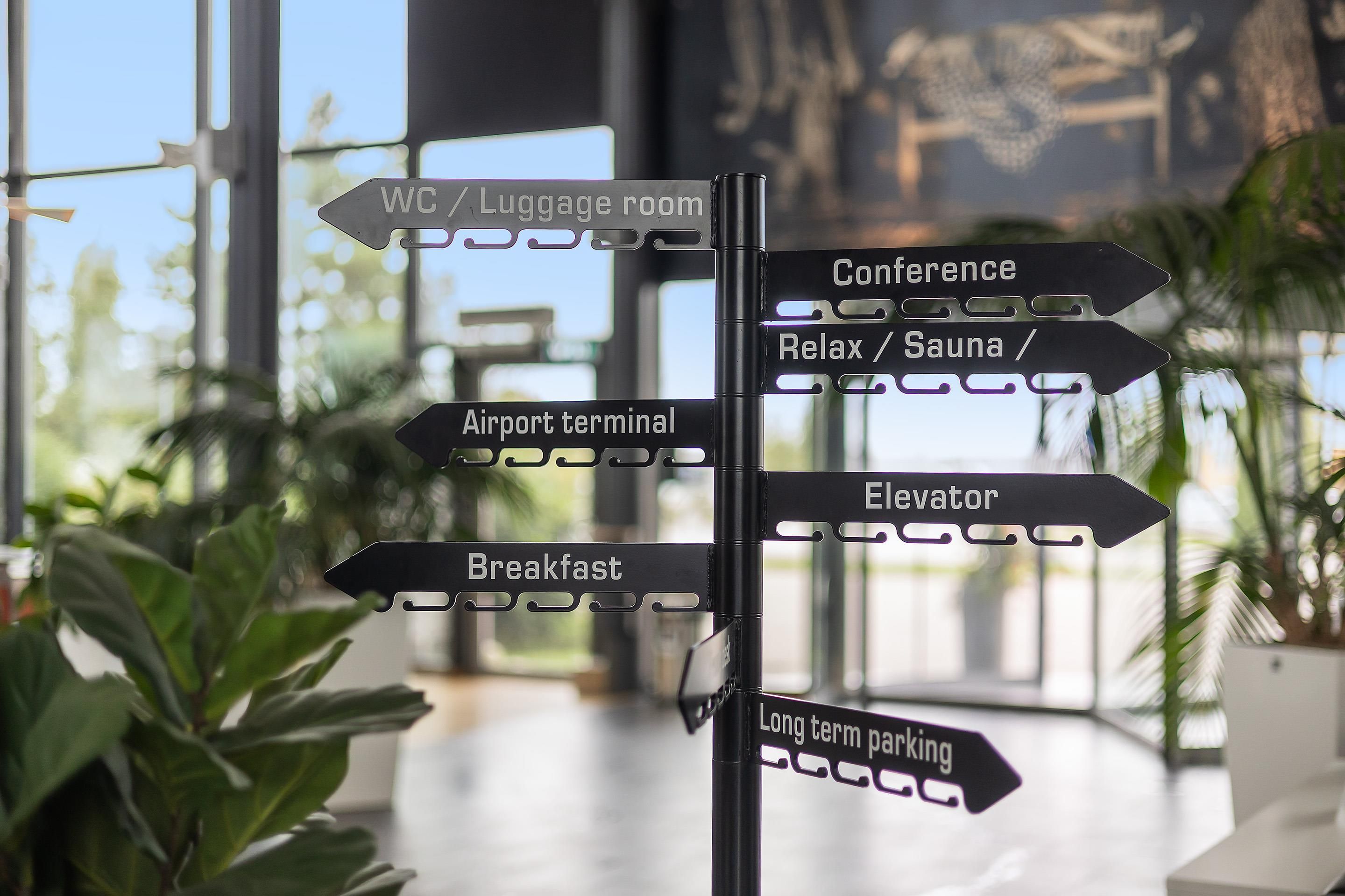 A directional signpost with multiple arrows pointing to various amenities such as WC, Luggage room, Conference, Relax/Sauna, Airport terminal, Elevator, Breakfast, and Long term parking.