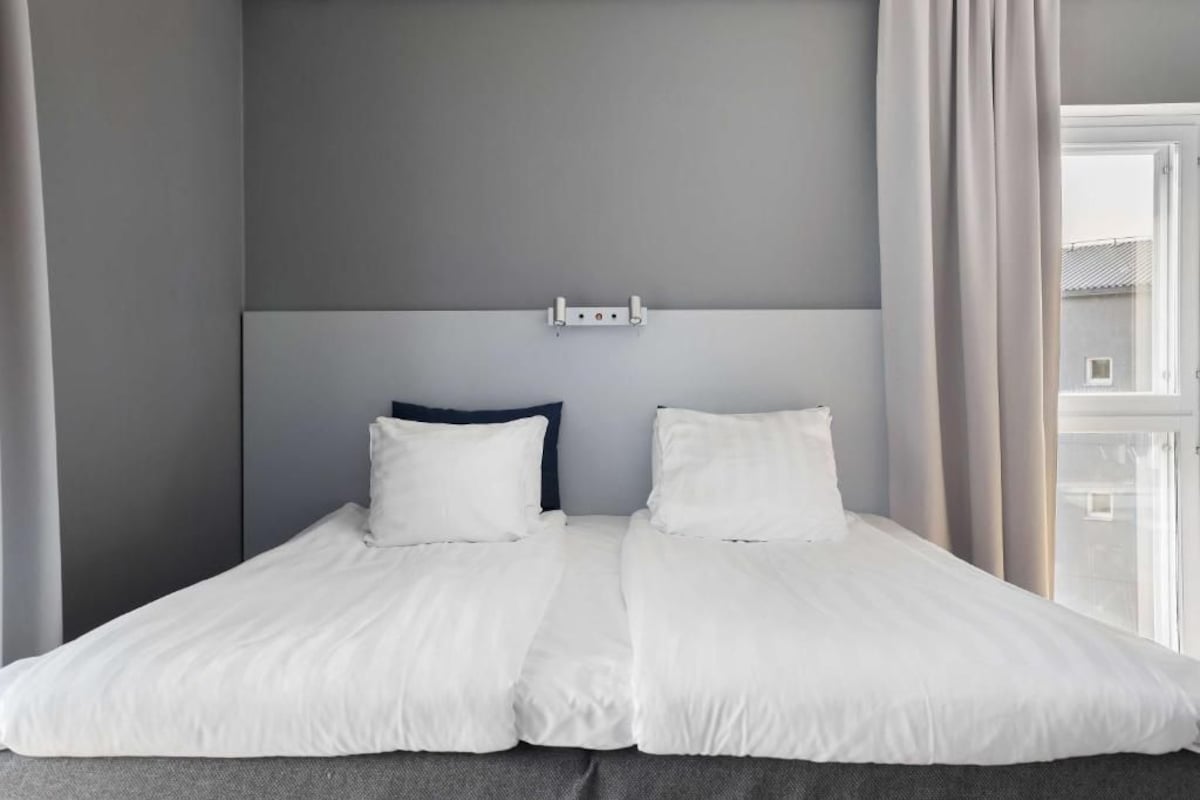 Uma cama de casal com roupa de cama branca, uma cabeceira escura e uma janela com cortinas.
