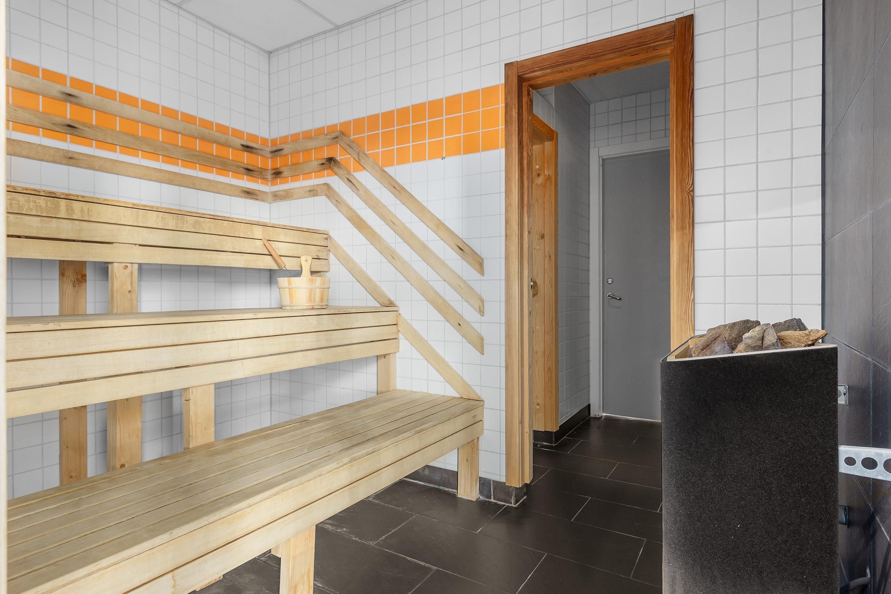 Una sala de sauna con bancos de madera y un banco de piedra, una pared con azulejos y detalles anaranjados, y una puerta que da a otra habitación.