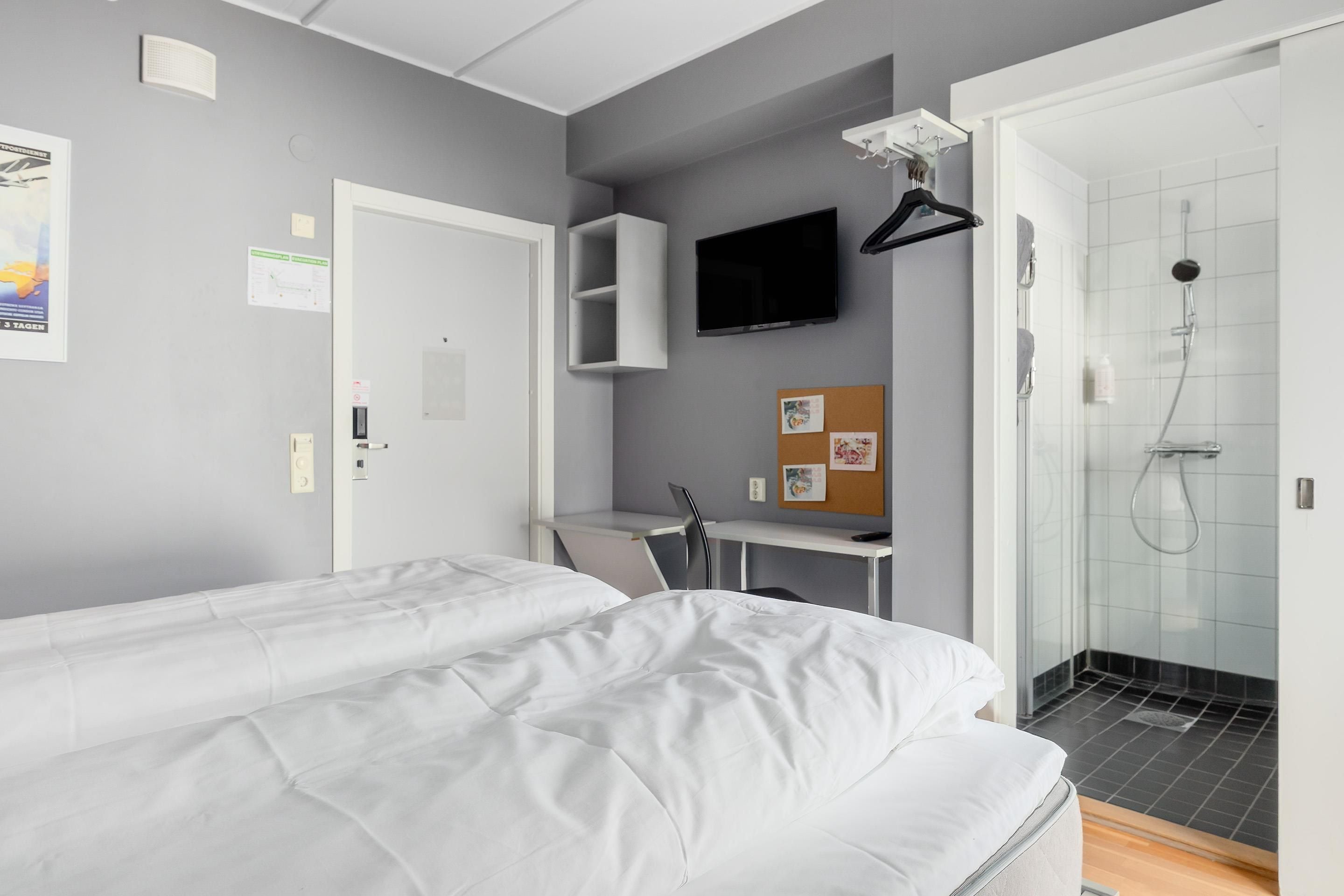 A hotel room with two single beds, a desk with a chair, a television, a shower, and a corkboard with pictures.
