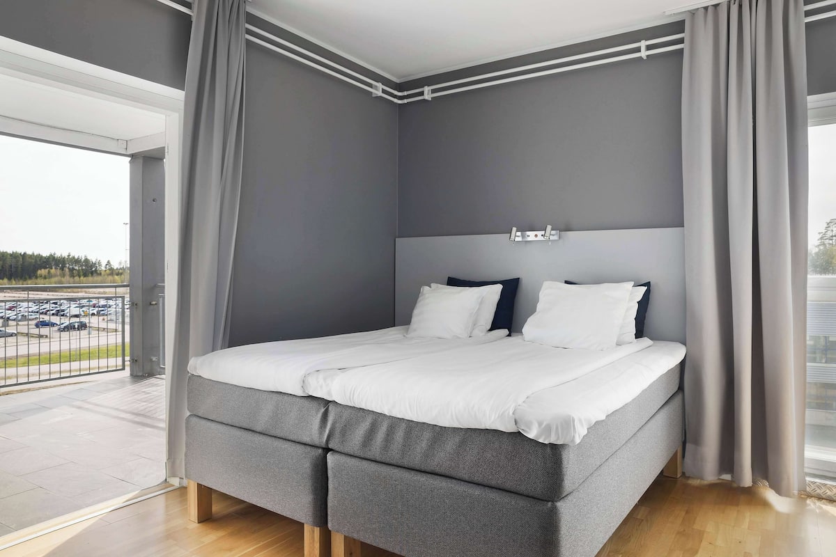 A modern bedroom with a large bed, grey walls, and a view of a parking lot through an open door.