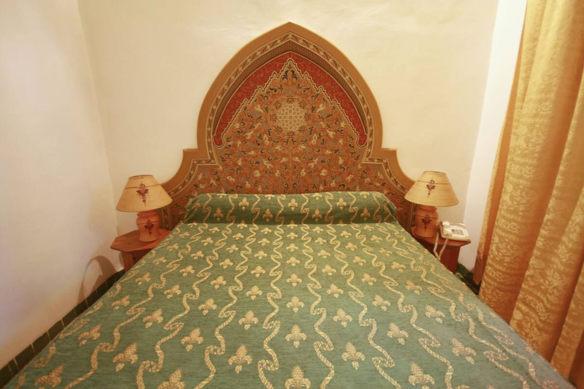 A room with a bed featuring a green patterned bedspread, a decorative headboard, two bedside lamps, and a telephone on a nightstand.