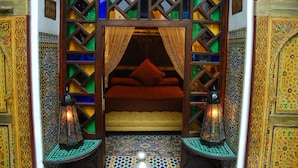 A traditional Moroccan room with a bed, ornate wooden lattice, patterned floor, and decorative lanterns.
