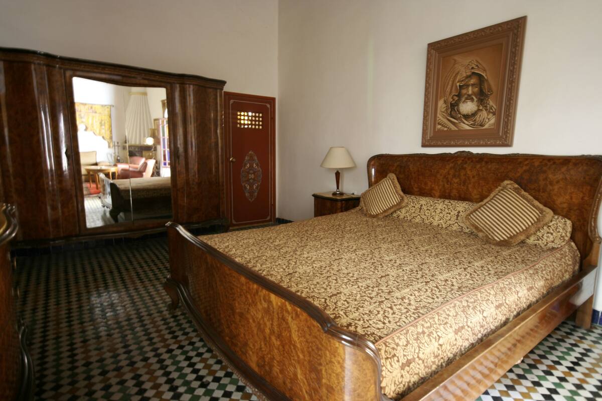 A bedroom with a wooden bed, a mirror, a framed picture on the wall, and checkered floor tiles.