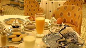A dining area with tables set for breakfast, featuring plates of pastries, a glass of orange juice, and a teapot.