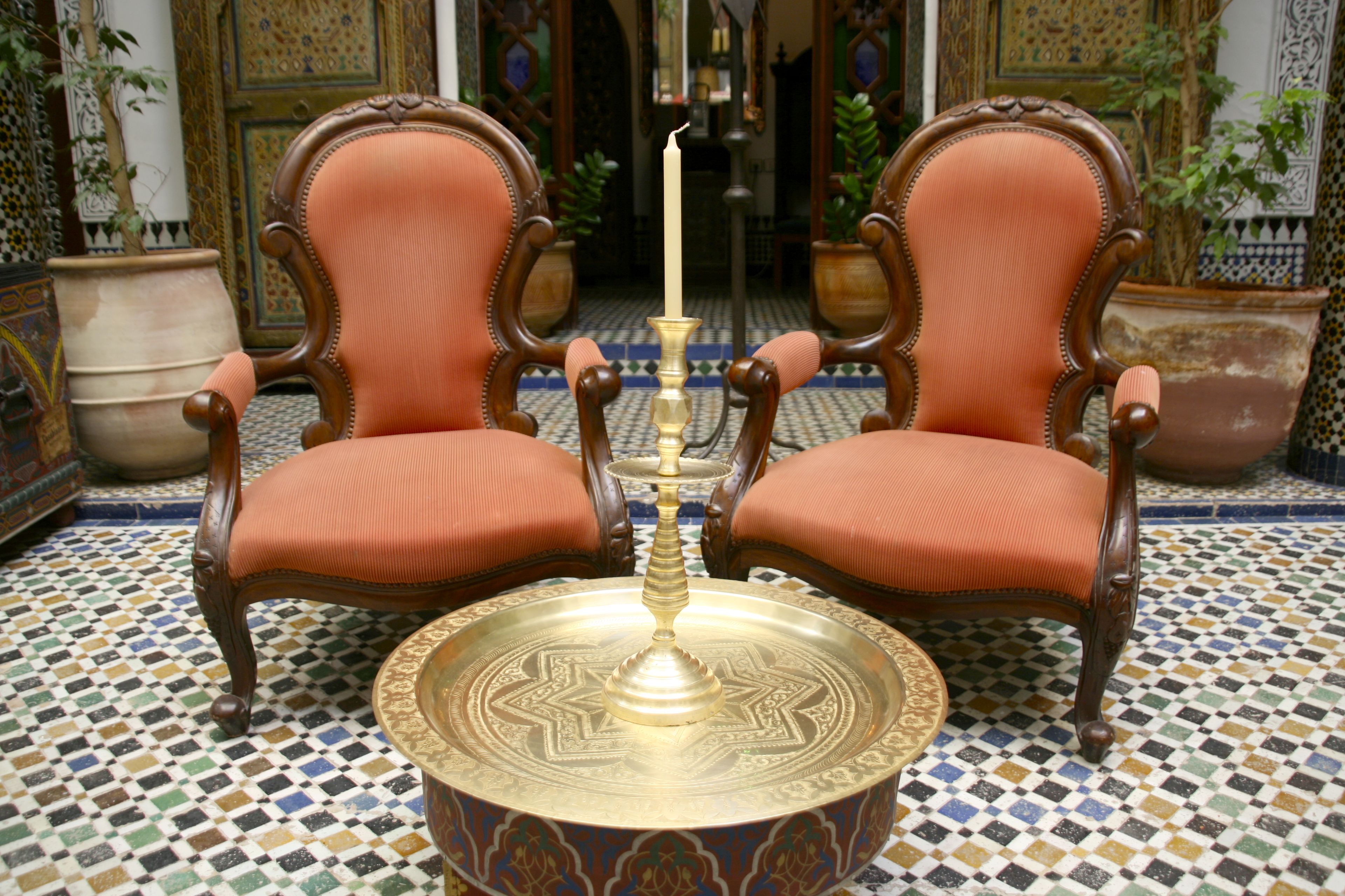 Duas cadeiras ornamentadas com assentos e encostos acolchoados, uma bandeja decorativa com uma vela no centro e um chão estampado.