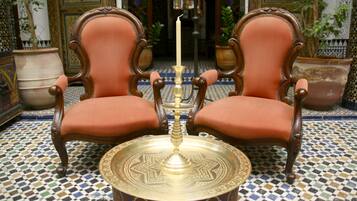 Duas cadeiras ornamentadas com assentos e encostos acolchoados, uma bandeja decorativa com uma vela no centro e um chão estampado.