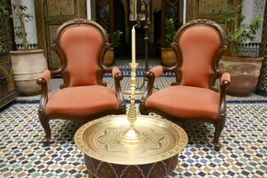 Duas cadeiras ornamentadas com assentos e encostos acolchoados, uma bandeja decorativa com uma vela no centro e um chão estampado.