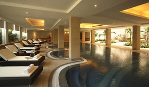 A large indoor swimming pool with lounge chairs and sunbeds, surrounded by pillars and potted plants. - Ellaa Hotel Gachibowli (Hyderabad)