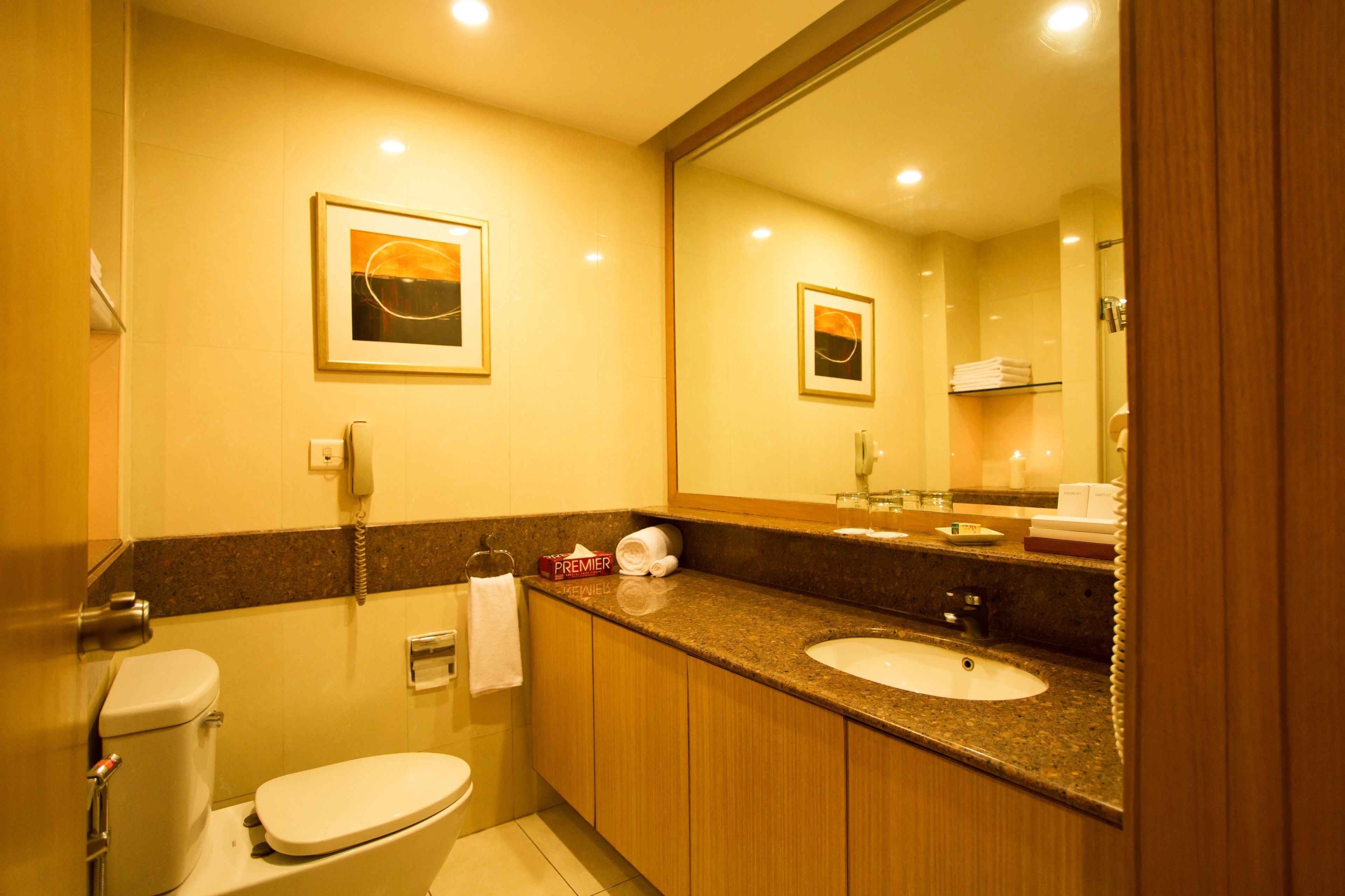 Casa de banho de hotel com sanita, lavatório e espelho. Encontra-se um telefone, uma toalha e uma caixa de lenços de papel sobre o balcão.