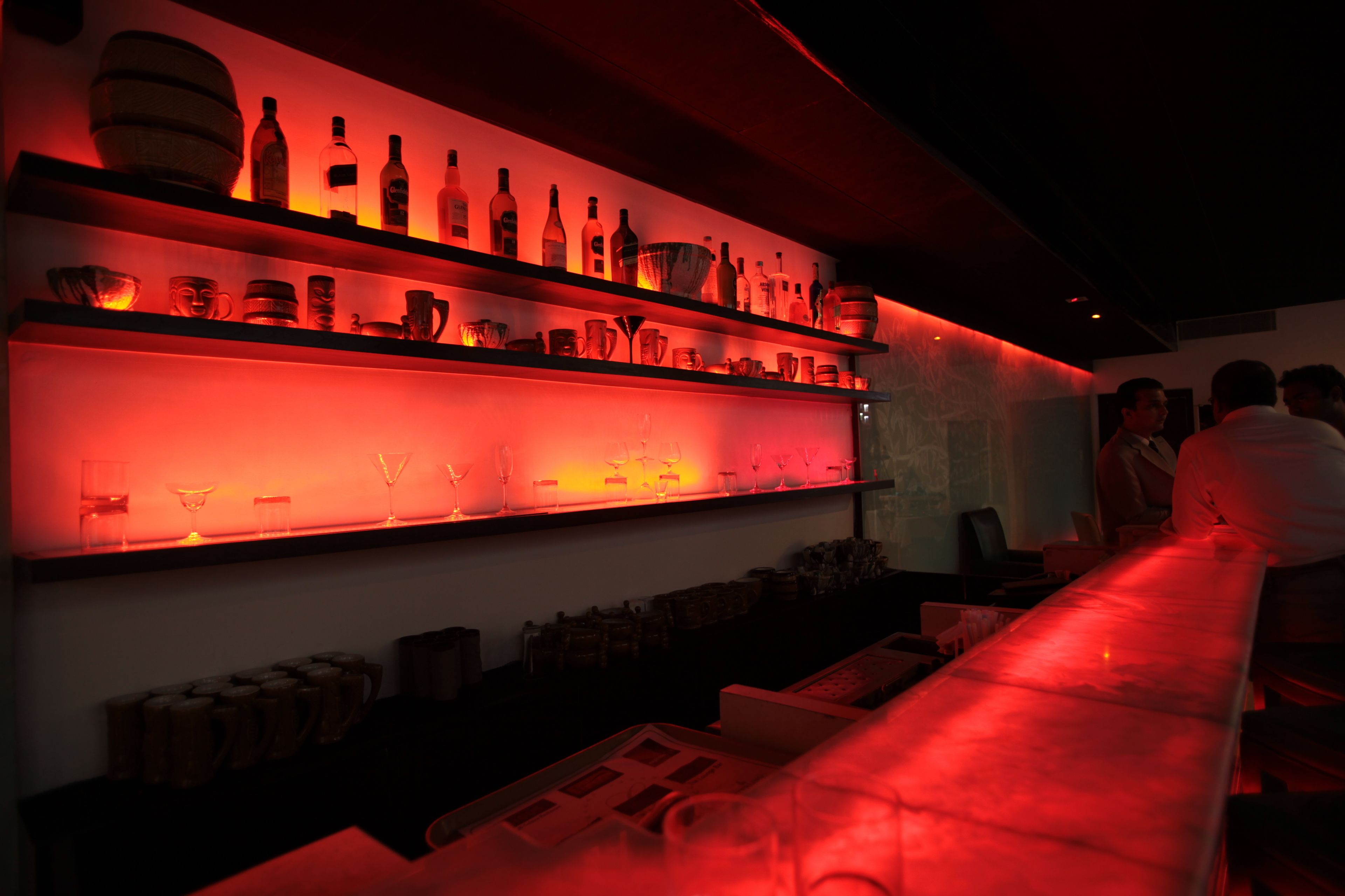 A bar area with shelves of bottles and glasses, a long counter, and a few people in the background.