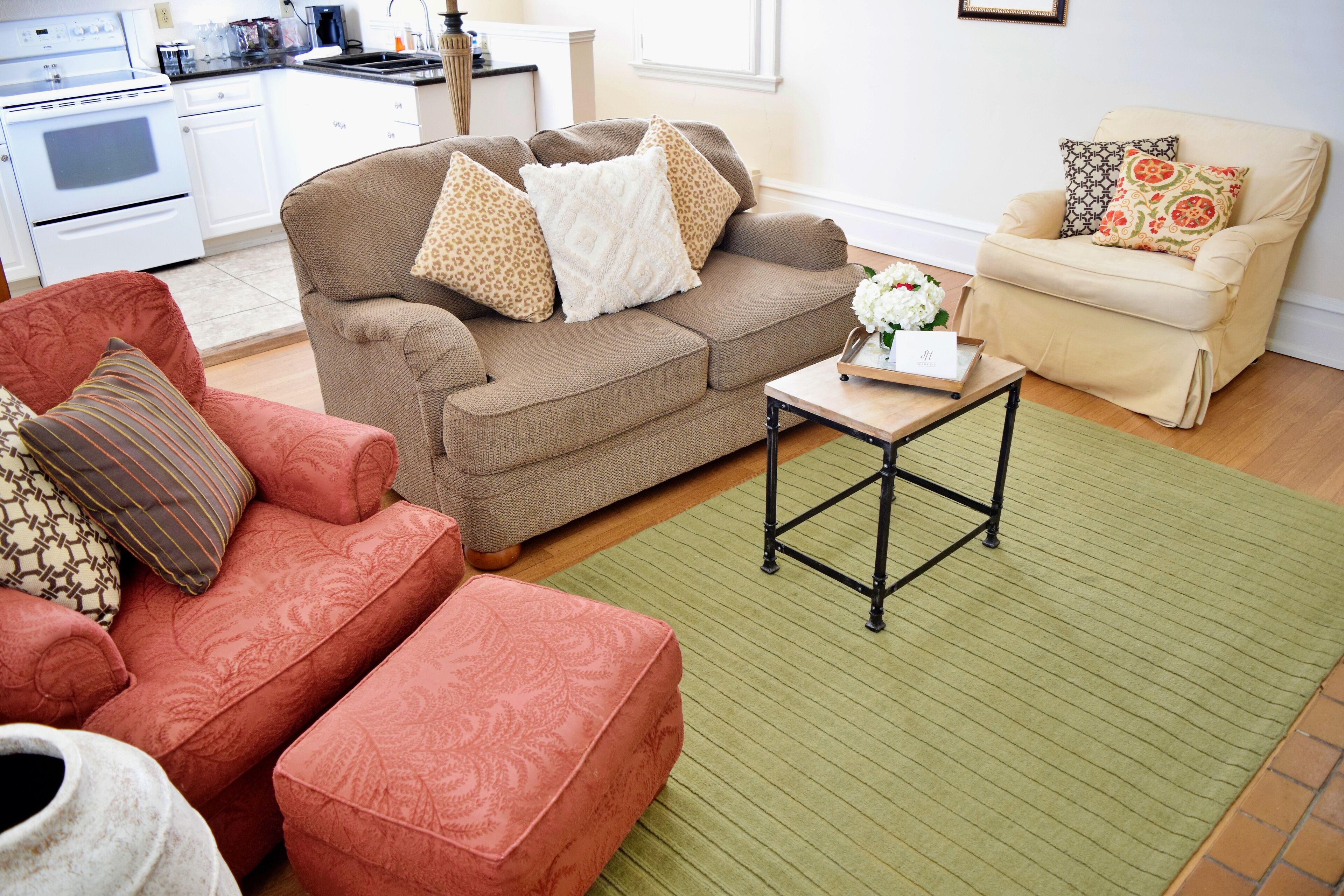 Ein Wohnzimmer mit einem braunen Sofa, einem roten Sessel und einem beigen Sessel. Ein kleiner Holztisch mit einem Blumenstrauß steht dort. Der Raum verfügt über einen grünen Teppich und einen Fliesenboden.