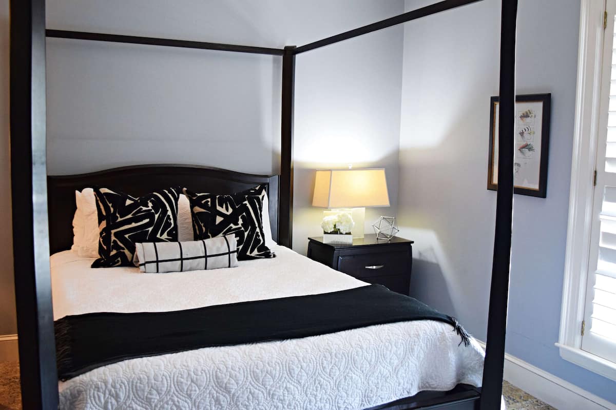 A four-poster bed with a black frame and white bedding, a bedside table with a lamp, and a framed picture on the wall.