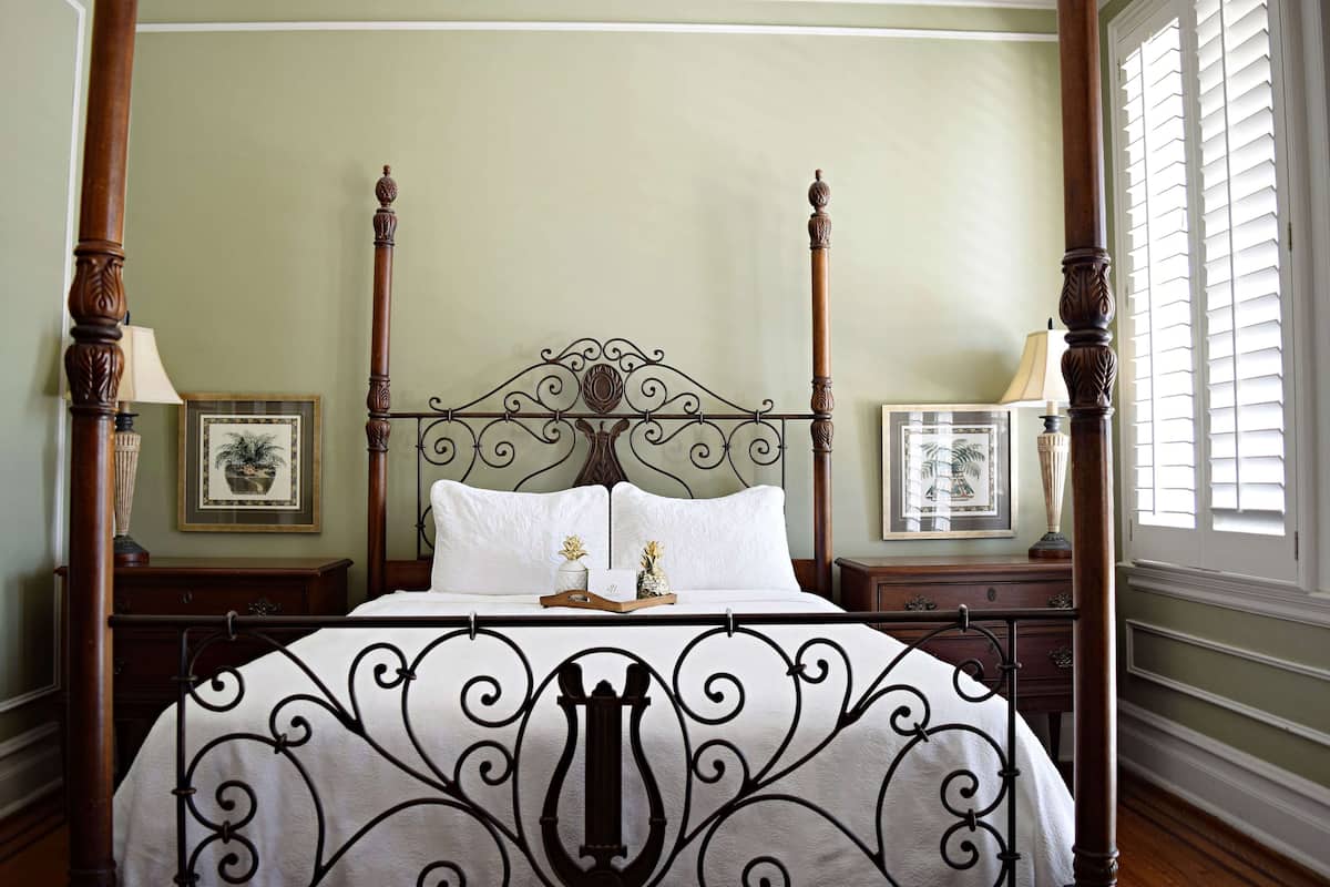 A four-poster bed with a white bedspread and a decorative metal headboard. There are two nightstands with lamps and framed pictures on either side of the bed.