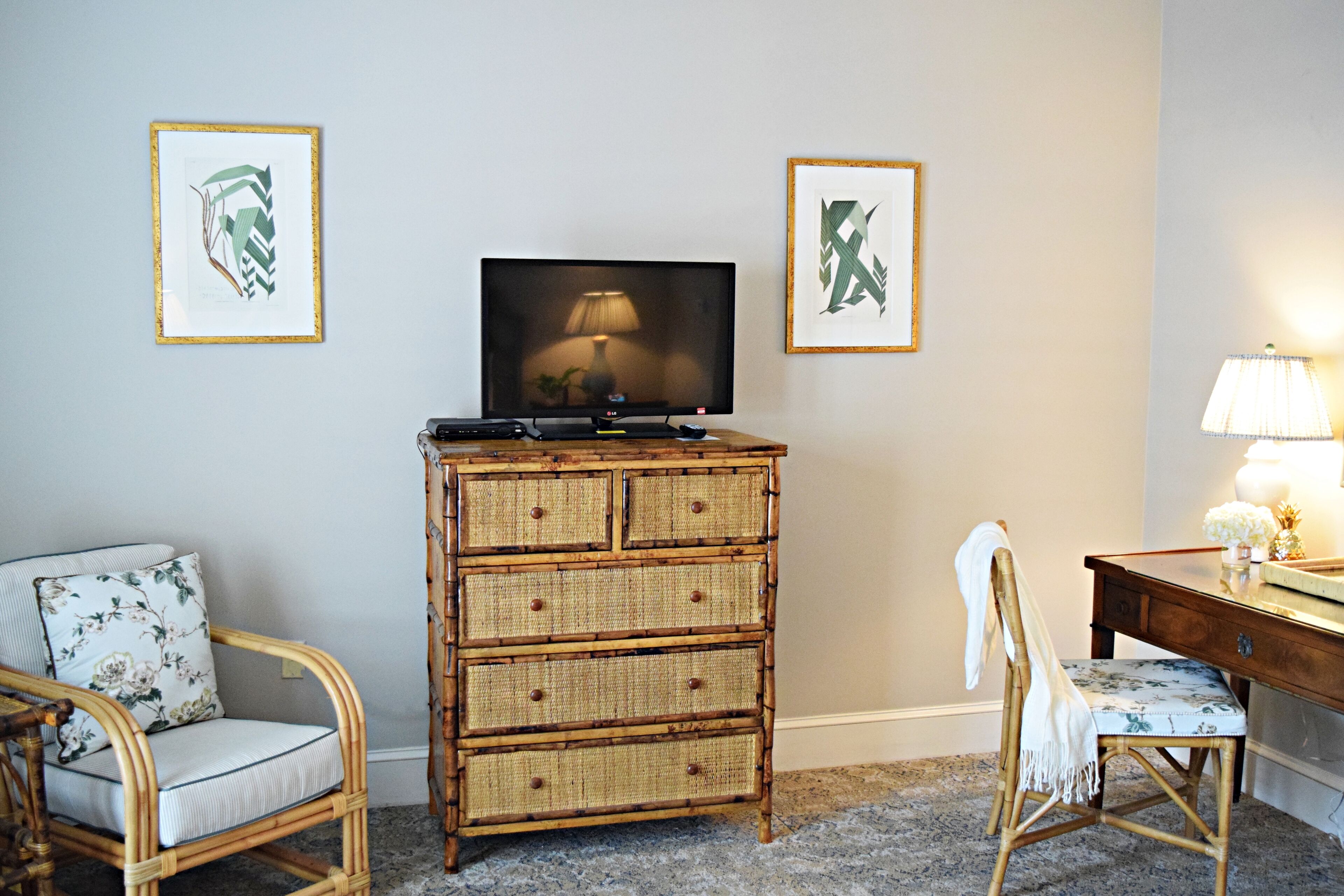 Une pièce comprenant un téléviseur posé sur un meuble, une commode en osier, deux chaises en osier et un bureau avec une lampe.